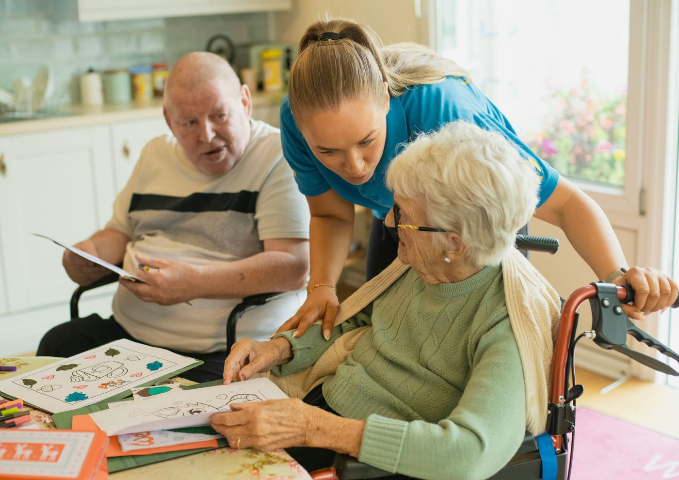 A care worker leaning in to chat with two older adults at a kitchen table